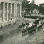 Image: lines of men in military uniforms plus others on horseback gather outside a large stone building with wide steps leading to its entrance and decorative columns along the entire facade.