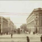 Image: a sepia photograph of a busy street, filled with horse drawn vehicles and pedestrians in 1870s attire, and lined with two and three storey stone buildings.