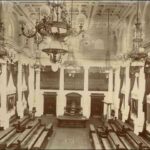 Image: a huge three storey high room with bench seating arranged in rows along the wall, facing inwards. There are long dark curtains draped above portraits, white columns along the walls, a decorative plaster ceiling and huge chandeliers