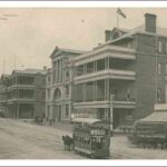 Image: a horse drawn tram travels down a wide dirt city street which is lined with a series of three and four storey buildings, many with balconies and verandahs, including a theatre with a large arched entrance and column decoration on the upper floors.