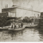 Image: Four men stand in a wooden row-boat next to landing steps on a riverbank. Two other empty row-boats are moored nearby, and a large building is visible on the shoreline in the immediate background