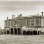 Image: a two storey stone building with a protruding arched portico flanked by verandahs. A clock sits centrally in a decorative parapet which partly conceals a low pitched roof.