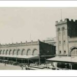 Image: Two large buildings with archway windows and a square-sided tower. Several cars of 1920s vintage are parked in front of the building