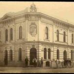 Image: sepia shot of building facade, with three men at the entrance, one by the side, and a horse-drawn carriage