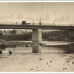 Image: a metal bridge with stone abutments and central pylon passes over a shallow river.