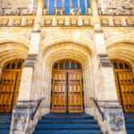 Image: The entrance to an ornate, historic building made of stone. Three large wooden doors are contained within corresponding stone archways