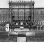 Image: a low stage, with the wall behind covered in decorative wooden panelling, supports rows of chairs, a small table and a lecturn. The stage also has further rows of chairs facing it.