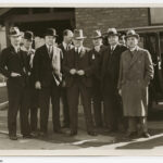 Image: A group of middle-aged Caucasian men dressed in early twentieth century suits and fedora hats stand in front of a brick building. A late-1920s model car is parked immediately behind the group