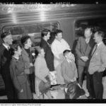 Image: Six people, both adults and children stand in front of a train on a station platform. They are laughing and conversing.