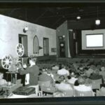 Image: A young man operates a film projector in a large church hall filled with seated people of all ages