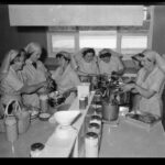 Image: A group of seven Caucasian women in 1960s attire that resemble nursing uniforms prepare and pack food in a small kitchen