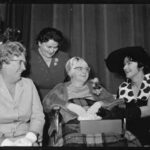 Image: A middle-aged Caucasian woman sits in a wheelchair and is wearing a fur stole and horn-rimmed glasses. Around her are seated three younger Caucasian women in formal dress