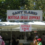 Image: A group of people patronise a food concession housed in a white tent. A sign in Italian on the tent reads “Sant’ Ilario, Bottega Delle Zeppole”