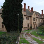 Image: Oblique view of the front of a large, two-storey stone mansion flanked by gardens