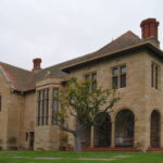 Image: A large, two-storey stone mansion bounded by a grass lawn. A small tree is visible next to the right foreground of the house