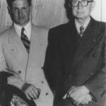 Image: Two clean-shaven Caucasian men dressed in mid-twentieth century suits pose for a photograph. The man standing at left is young and has dark hair. The other, also standing, is elderly with white hair and is wearing thick-framed glasses