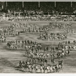 Image: An oval and grandstands filled with people, children dance on the oval in circular formations