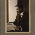 Image: A photographic portrait of a man in a suit sitting at a desk and examining a book