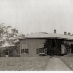 Image: A path leading through lawn to Cummins House at Novar Gardens. The house is constructed of red bricks. It has a bay windows and shutters on all windows