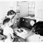 Image: Two adult women and a young boy sit in front of a large two-way radio receiver. One woman holds the radio's transmitter in her hand. The boy has an open textbook in front of him