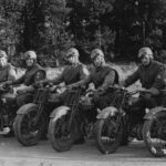 Image: Nine men wearing Second World War-era military uniforms each sits astride a motorcycle. Each man is wearing a helmet with goggles, and all are arranged in a line abreast of one another