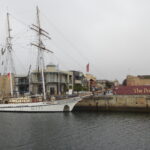 Image: A two-masted sailing ship is tied up alongside a long concrete wharf. A mix of old and modern buildings are visible in the background