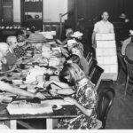 Image: A group of women sit on either side of a long table arranging paper documents, a man walks by with a trolley stacked with papers.