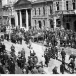 Image: A large group of men stand in the street looking towards the front of a building.