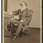 Image: A man wearing a suit sitting in a chair with one hand resting on a small pedastal