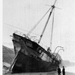 Image: A large sailing ship sits stranded on the beach, two men stand observing from the side