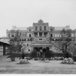 Image: Front view of a historic building with a small garden and trees at the front