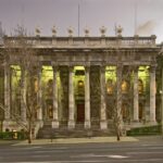 Image: a large stone building with two storey high columns along the entire facade, and wide stone steps leading up to its front entrance, is lit up with green lights at dusk.