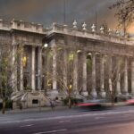 Image: A large stone building with two storey high columns lining its front is lit up by green lights at dusk.