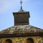 Image: Modern photograph of a bluestone building with arched windows surrounded in red brick and a rusty tin roof, topped with a weather vane.