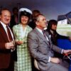 Image: a man in a grey suit sits at a wheel steering a small boat. Behind him other men and a woman in a green dress and captain's hat hold champagne and smile at the camera.