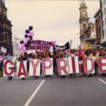 Image: men and women in 1970s era clothes march down a city street with pink and black balloons and signs reading "gay pride" and "homosexual liberation"