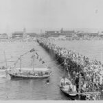 Image: Crowds of people line the Glenelg Jetty to for Proclamation Day celebrations. A small yacht sits in the water perpendicular to the jetty