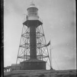 Image: A metal lighthouse stands on a desolate island. Two men stand in the lantern room at the top of the lighthouse, while a third sits outside the keepers’ quarters at its base. A horse and cart stands next to the seated man