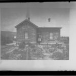 Image: A man stands in the front doorway of a modest-sized stone villa. A small flower garden is located in front of the house