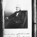 Image: A lithograph of a middle-aged man in Victorian attire seated at a table and holding a book