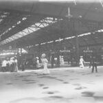 Image: the interior of a railway station with at least three platforms visible. The curved roof features skylights and is supported by metal beams. In the foreground are a number of men and women in early 20th century clothing.