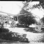 Image: A large stone house bordered on one side by a well-tended garden. A woman stands on the verandah of the house