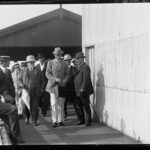 Image: A man in a hat and suit walks on a jetty ahead of a group of other men. The side of a corrugated metal building is visible to the right of the men