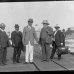 A man in a suit and hat is greeted by other men on a jetty. A coastal town is visible in the background