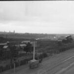 Image: a series of small fenced enclosures with a scattering of metal sheds line a dirt road with tramway.