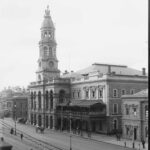 Image: A large stone building with a central tower capped by a dome. People cross a street in the foreground