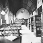 Image: A long, well-lit room containing several shelves of books on one side and six reading tables on the other