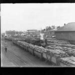 Image: Several train cars full of sacks are parked on a long wharf. A steam locomotive is positioned within the midst of the train cars. Three large stone buildings are visible in the background