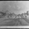 Image: Two and three storey residences and commercial buildings are set back from a wide tree lined dirt street upon which pedestrians and horse-drawn vehicles travel.