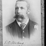 Image: A photographic head-and-shoulders portrait of a young Caucasian man wearing a late-Victorian era suit and sporting a very large handlebar moustache